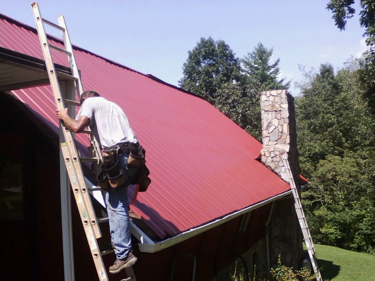 Roofer on a ladder beside a red metal roof and stone chimney — Soffit & Fascia Repair by Roofing Brothers in Torrance
